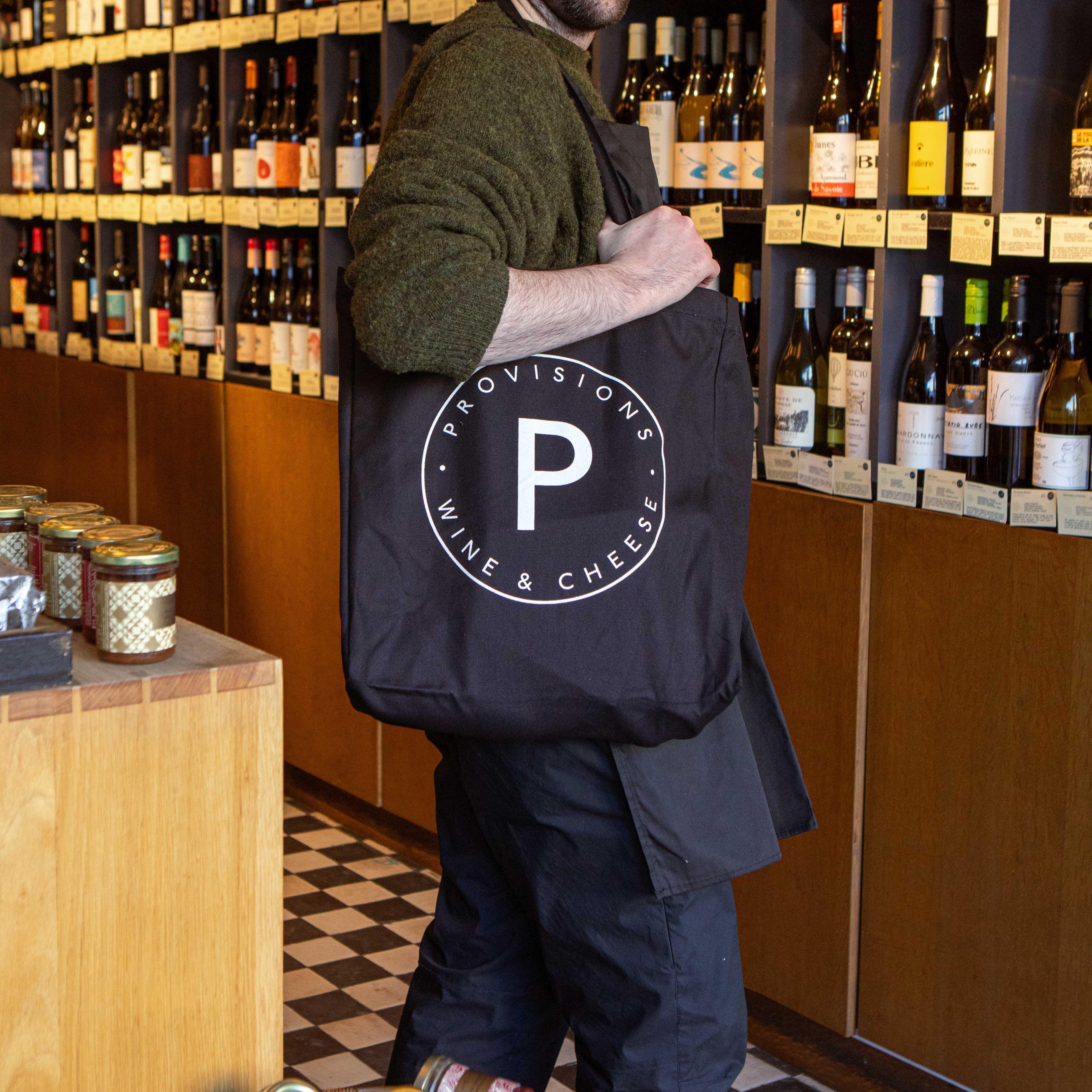 Man holding a black tote bag with 'Provisions Wine & Cheese' logo in a wine & cheese store.